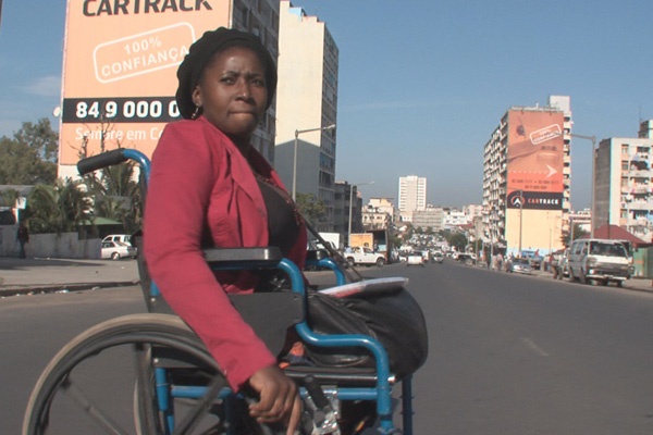 Uma mulher negra em uma cadeira de rodas no meio de uma avenida larga com piso de asfalto. Veste blusa e calças pretas e casaco cor de vinho. Ao fundo, prédios altos, automóveis e o céu azul.