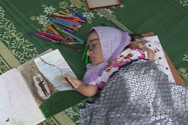 Uma mulher muito pequena está deitada sobre um tapete verde. Usa lenço na cabeça e está coberta com um tecido prateado. Ela faz um desenho com lápis de cor. 