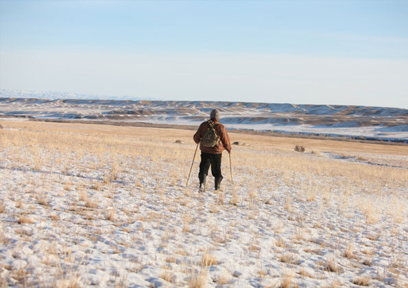 Uma pessoa, no centro da foto, caminha sobre planície arenosa. Está de costas e segura bengalas. Usa roupas de frio com calça, casaco e gorro.