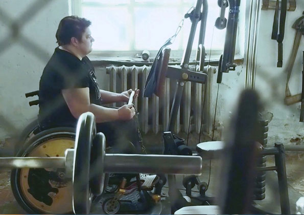 Uma mulher branca corpulenta de cabelos curtos está em um local rústico com aparelhos de musculação. Ela está em uma cadeira de rodas segurando com as duas mãos uma barra de um dos aparelhos.