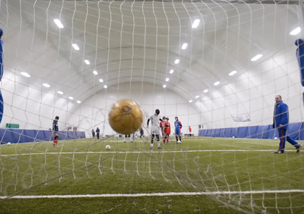Ponto de vista de atrás de uma goleira no exato momento que a bola amarela de futebol encosta na rede ainda no ar. Ao fundo, jogadores em um campo de grama sintética. 