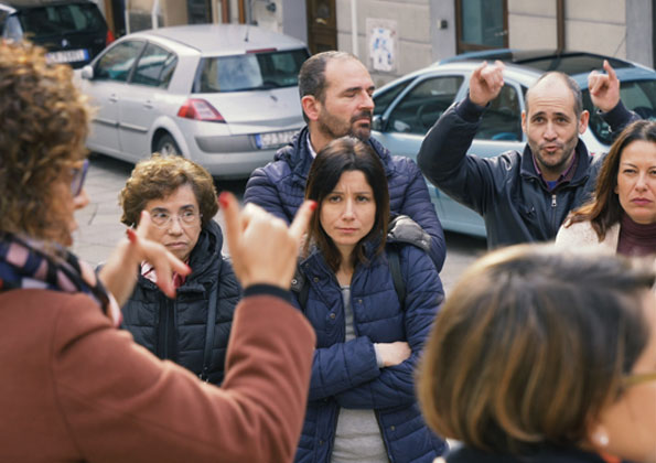 4 mulheres e 2 homens  estão próximos a uma intérprete de língua de sinais que está de costas, no lado esquerdo. Um homem a direita, está com os braços levantados sinalizando para a intérprete.