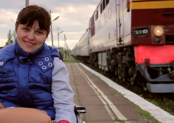 Jovem branca sentada em uma cadeira de rodas sorrindo em uma estação de trem ao ar livre. No lado esquerdo há um trem cor de vinho.
