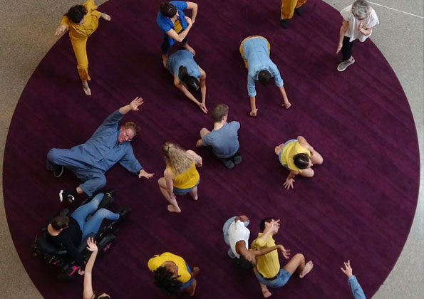 Imagem vista do alto de um grupo de pessoas que dançam sobre um tapete redondo roxo. Usam roupas amarelas e azuis. Há um homem em uma cadeira de rodas. Estão em poses diferentes.