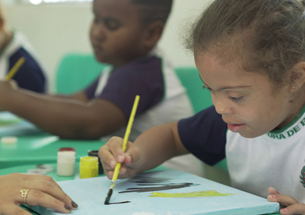 Em 1º plano, uma menina com síndrome de down pinta com pincel uma tela que está sobre uma mesa em ambiente escolar. A mão de uma mulher segura a tela. Ao lado dela,  dois meninos desfocados.