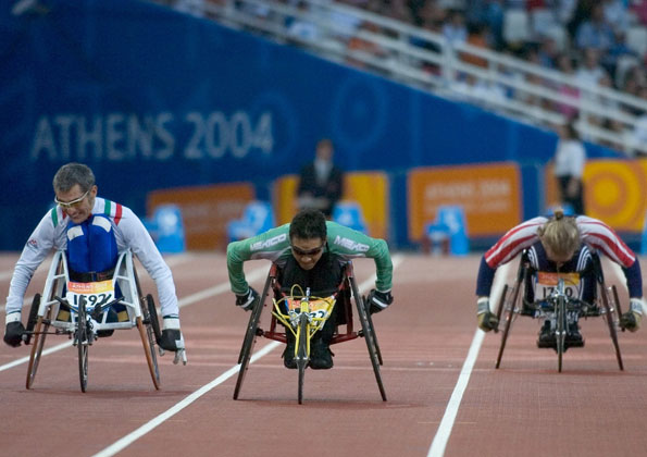 Em uma pista de atletismo, 3 atletas com cadeiras de rodas competem. Seus troncos estão para frente encostados em suas pernas flexionadas. Escrito ao fundo “Atenas 2004”.