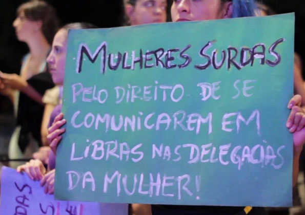 Uma mulher segura um cartaz verde com os dizeres “Mulheres Surdas pelo direito de se comunicarem em libras nas delegacias da mulher!” Atrás dela há outras mulheres.
