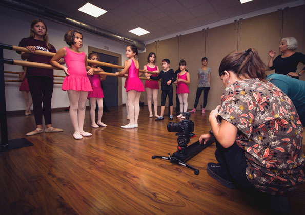 Em uma sala de dança, seis meninas e um menino seguram uma barra de madeira. Uma mulher está agachada na frente deles filmando. Outra observa.