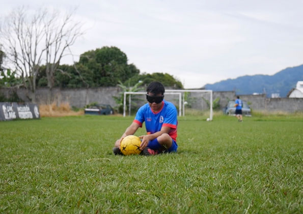 Um menino com um tapa olho sentado no meio de um campo de futebol com uma bola nas mãos. Mais atrás, outro rapaz joga. Ao fundo, um muro e montanhas.